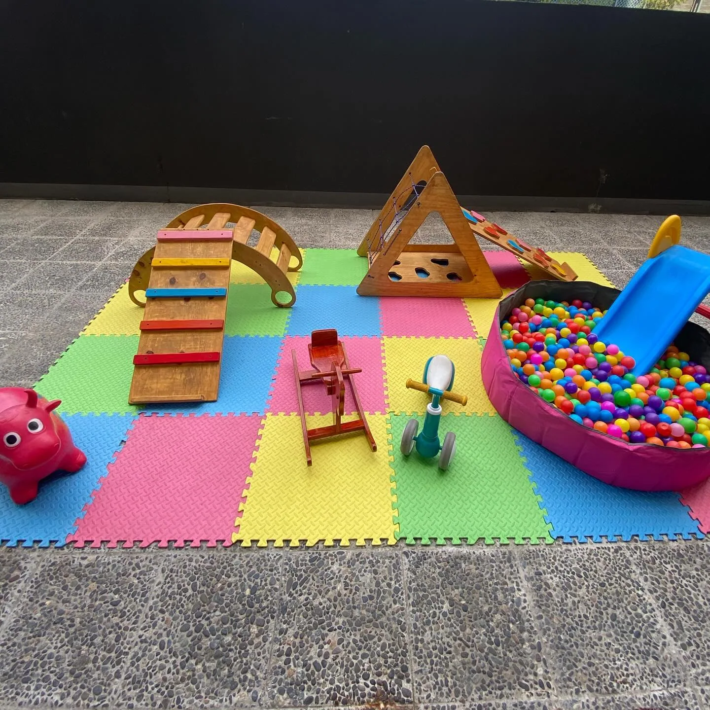 Montaje estilo Pikler en un patio de piedra: arco balancín de madera, triciclos y piscina de pelotas