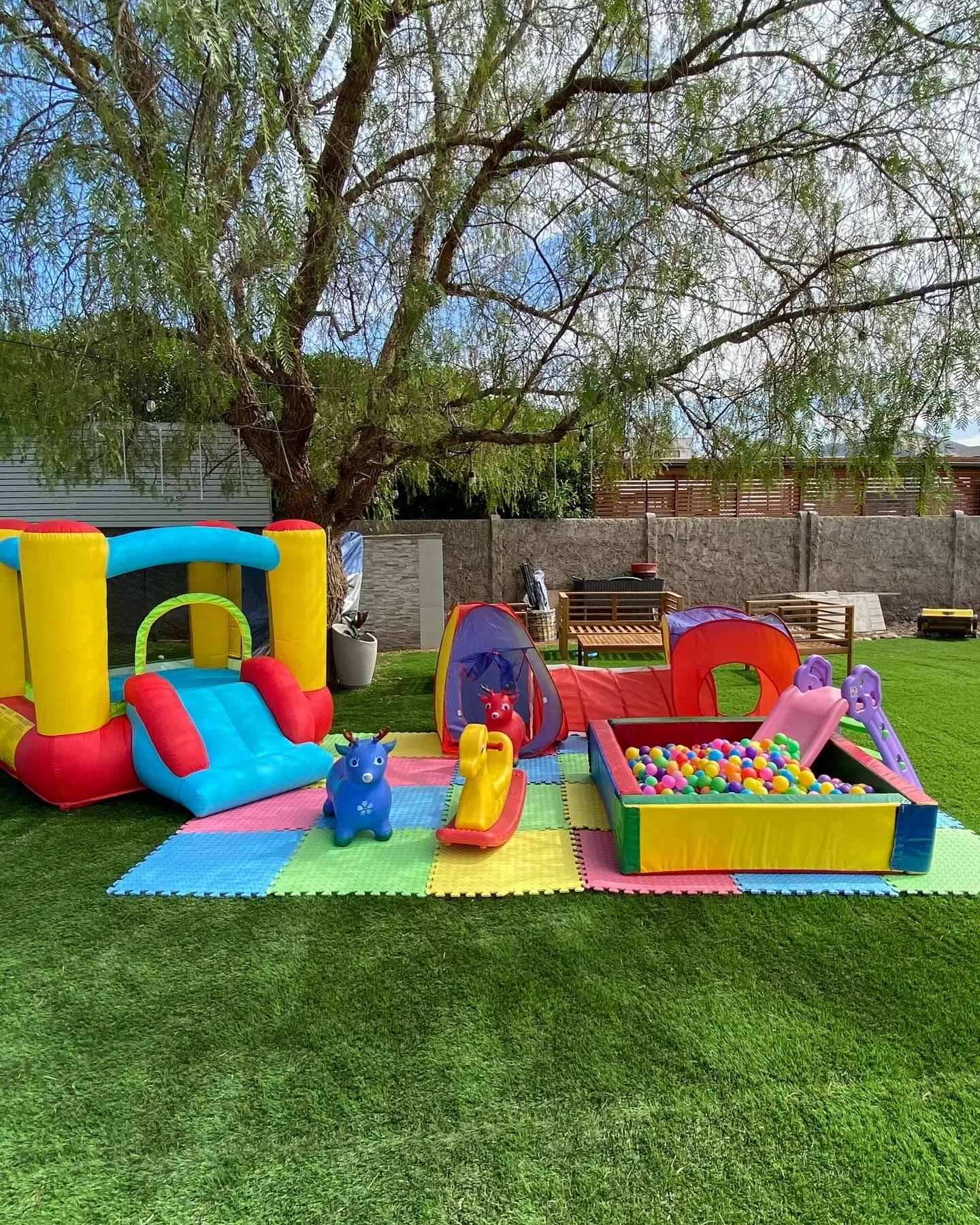 Castillo inflable chico con resbalín bajo un árbol grande, sobre pasto sintético